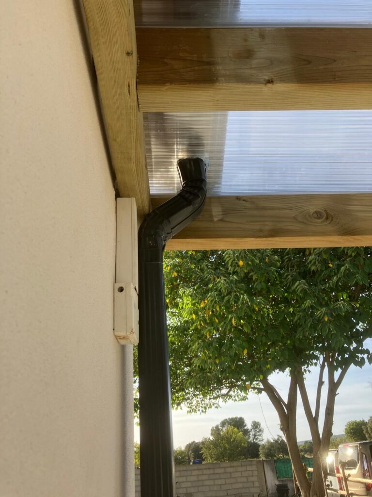 Gouttière fixée sous toit en bois, arbre en arrière-plan.